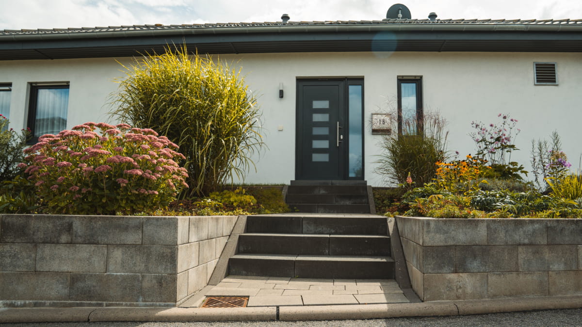 Gepflasterter Eingangsbereich mit breiter Treppe, Stützmauern und bepflanztem Vorgarten vor einem Wohnhaus.