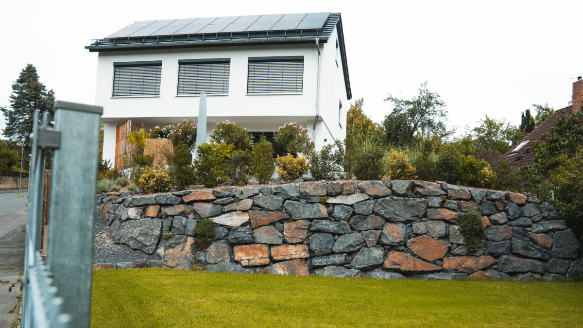 Natursteinmauer aus grau-braunen Bruchsteinen vor einem modernen Wohnhaus mit Garten.