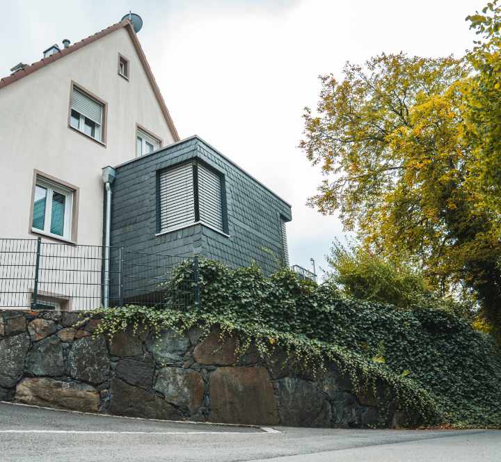 Natursteinmauer aus dunklen Basaltsteinen mit dichter Efeubepflanzung vor einem Wohnhaus.