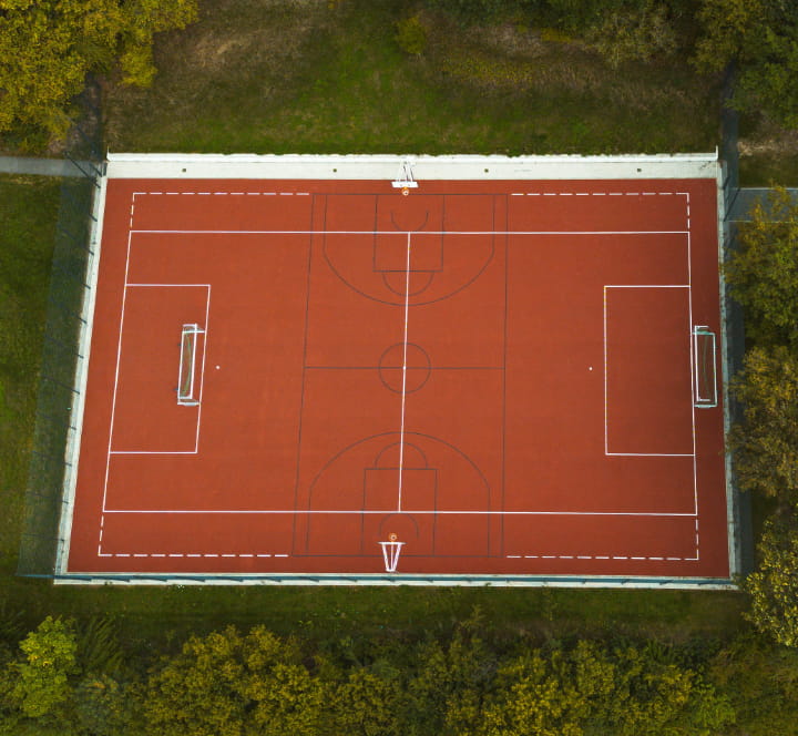 Luftaufnahme eines roten Multifunktions-Sportplatzes mit Fußballtoren und Spielfeldmarkierungen an der Jugendbildungsstätte Neukirchen.