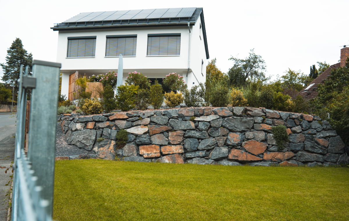 Natursteinmauer als Grundstückseinfassung vor einem modernen Wohnhaus mit Garten und Rasenfläche.