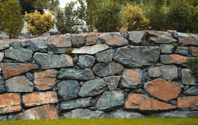 Natursteinmauer aus unregelmäßig gesetzten, grau-braunen Steinen mit Bepflanzung im Hintergrund.