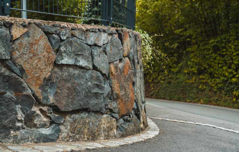 Natursteinmauer aus dunklen Basaltsteinen als Einfassung entlang einer Straße mit Zaun und bewaldetem Hang.