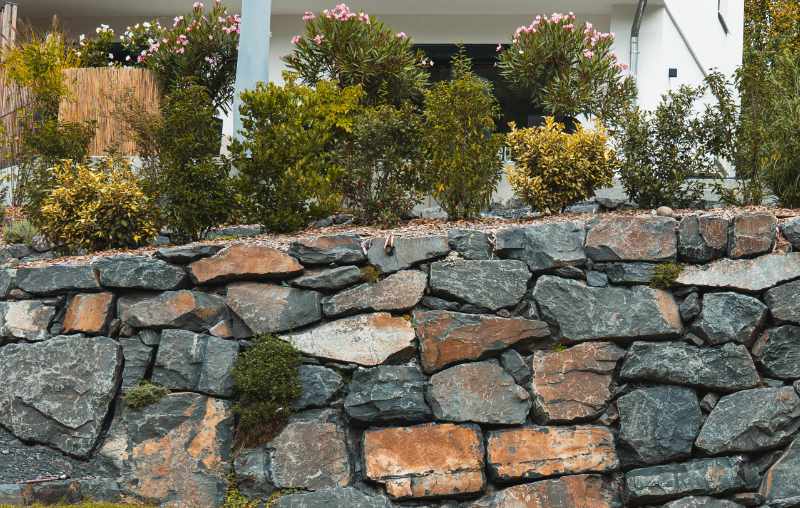 Nahaufnahme einer Natursteinmauer aus unregelmäßigen grau-braunen Steinen mit bepflanztem Garten darüber.