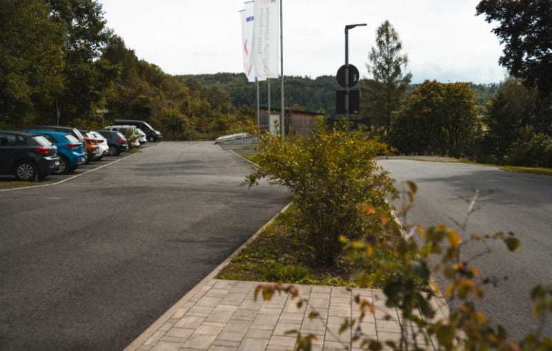 Asphaltierter Parkplatz mit Stellplätzen, Gehweg und bepflanzter Mittelinsel an der Jugendbildungsstätte Neukirchen.