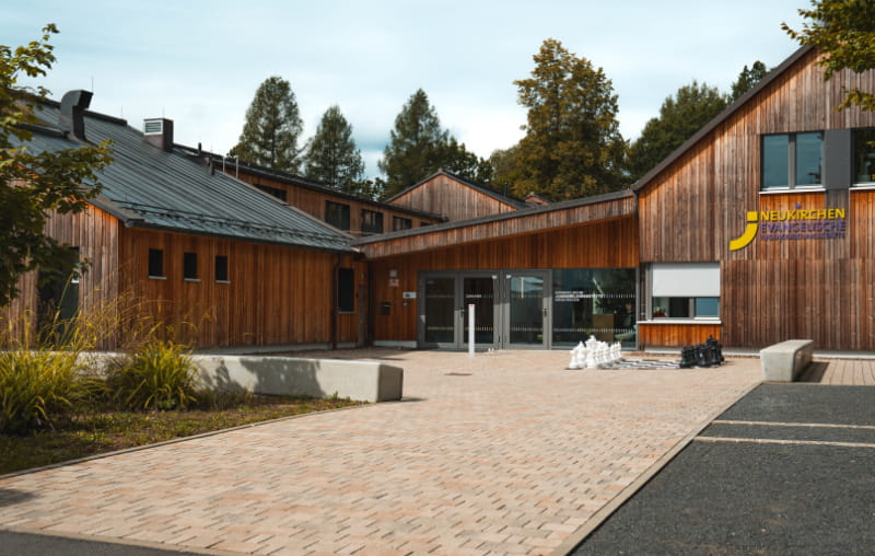 Gepflasterter Eingangsbereich der Evangelischen Jugendbildungsstätte Neukirchen mit Holzfassade und Sitzbänken.