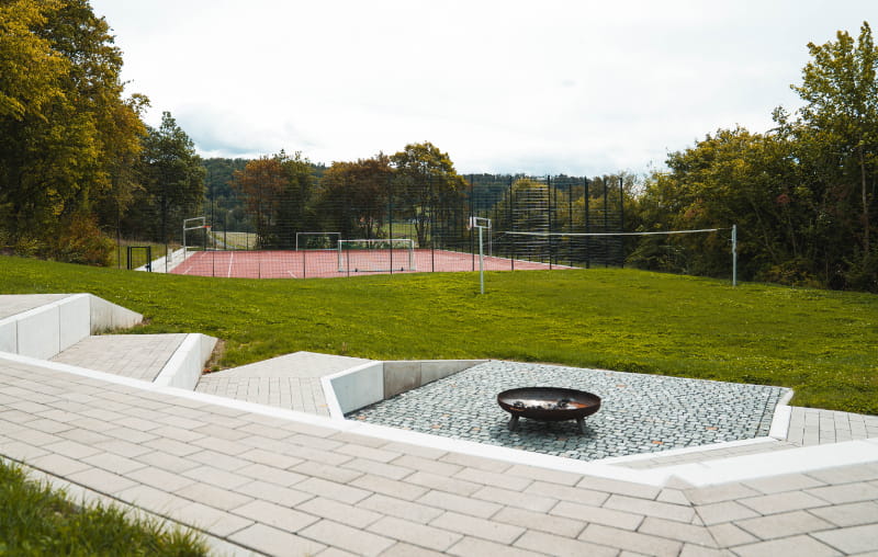 Gepflasterter Außenbereich mit Feuerstelle, Sitzstufen und Blick auf den Sportplatz der Jugendbildungsstätte Neukirchen.