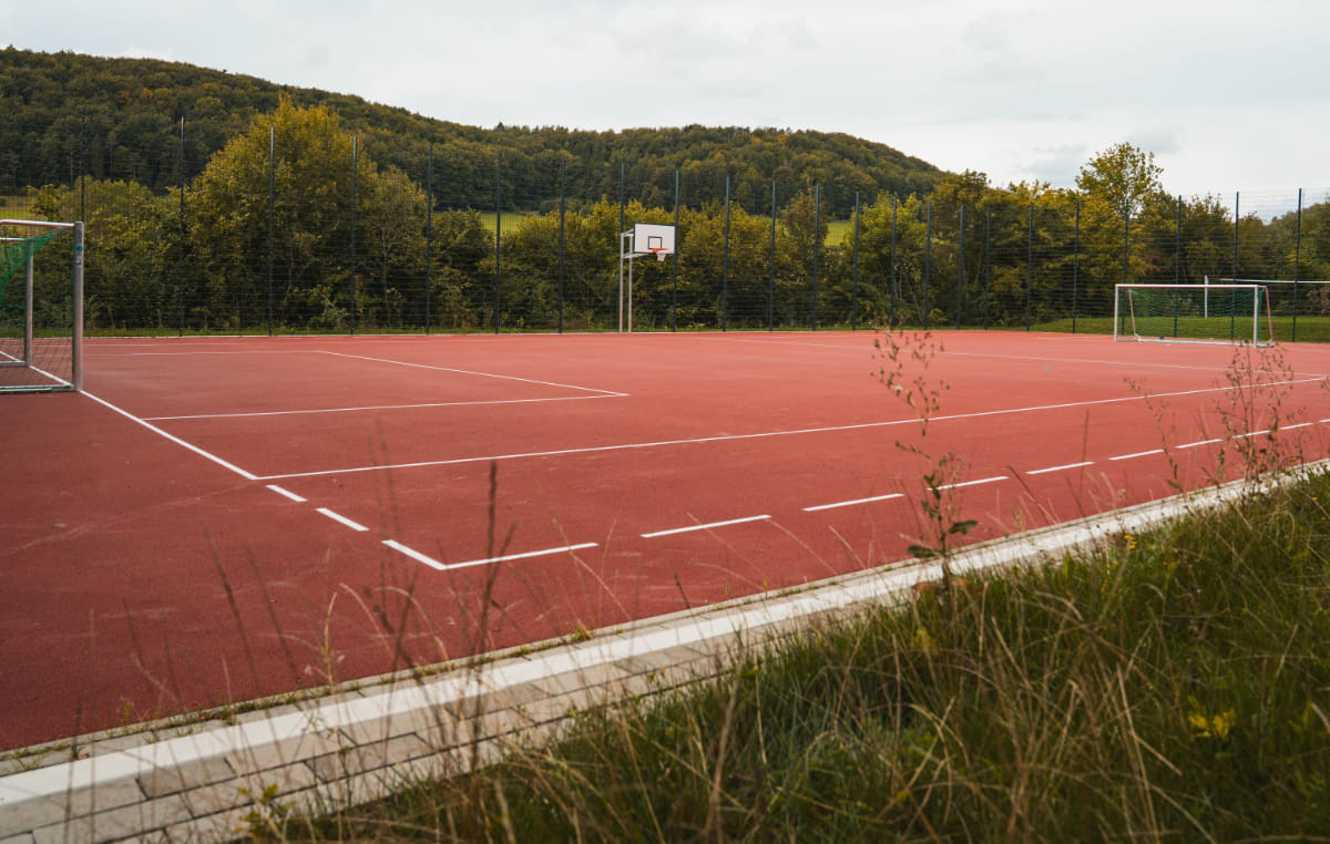 Roter Multifunktions-Sportplatz mit Fußballtoren und Basketballkorb in bewaldeter Umgebung an der Jugendbildungsstätte Neukirchen.