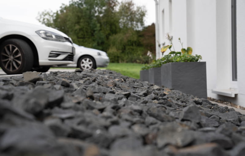 Schotterfläche mit Pflanzkübeln neben dem Gebäude und abgestellten Autos im Hintergrund.