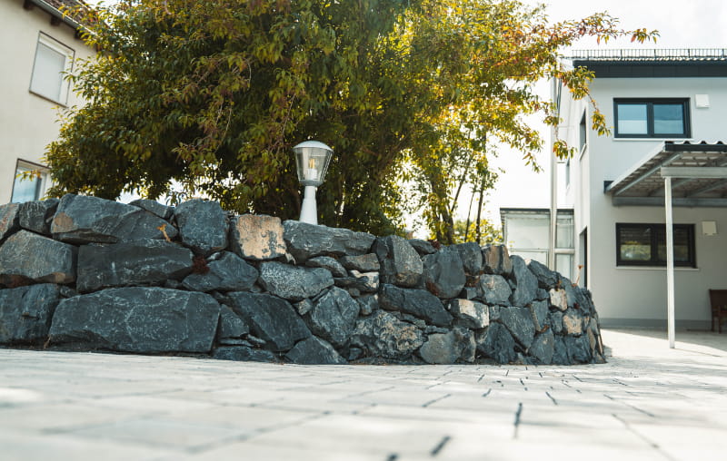 Gepflasterte Hoffläche mit geschwungener Natursteinmauer und Gartenbereich vor einem Wohnhaus.