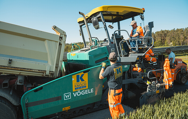 Straßenfertiger beim Einbau der Asphalt-Tragdeckschicht mit Anlieferung des Asphaltmischguts per LKW.